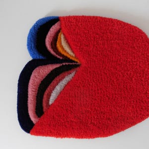 Op de afbeelding: Abstract tapijt met een getextureerd oppervlak. Het heeft een groot rood gedeelte en een gedeelte met gelaagde bogen in blauw, zwart, roze, wit en oranje. Ontworpen als decoratieve vloerbedekking voor een woonruimte.