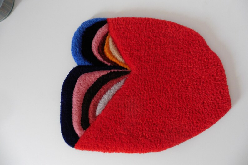 Op de afbeelding: Abstract tapijt met een getextureerd oppervlak. Het heeft een groot rood gedeelte en een gedeelte met gelaagde bogen in blauw, zwart, roze, wit en oranje. Ontworpen als decoratieve vloerbedekking voor een woonruimte.