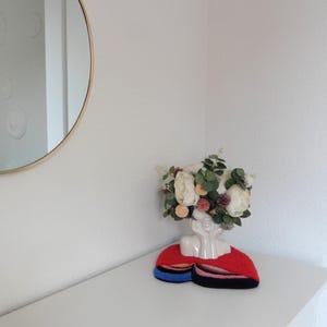 Op de afbeelding: Een witte keramische bustevase met een bloemstuk van witte en perzikbloemen, groene bladeren en kleine paarse accenten. De vaas staat op een rood, blauw en zwart viltstuk. Op de achtergrond een goudomrande ronde spiegel.