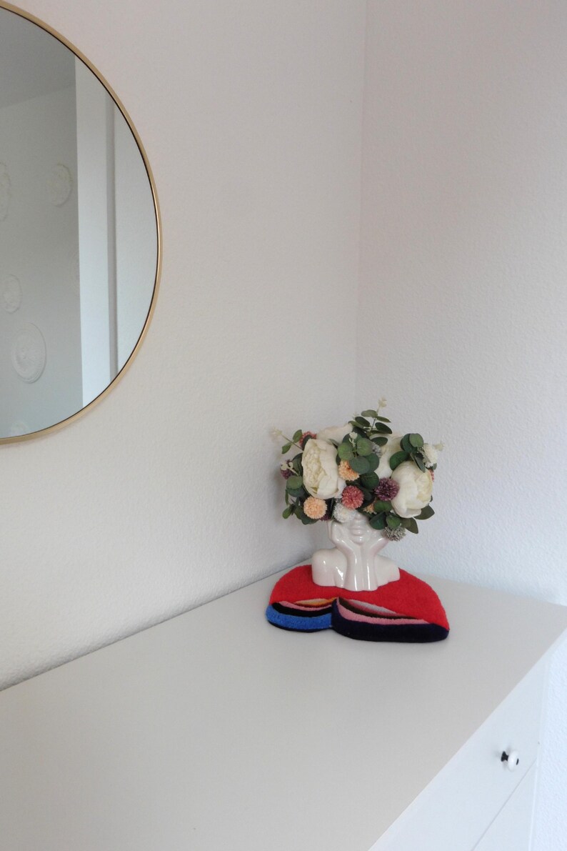 Op de afbeelding: Een witte keramische bustevase met een bloemstuk van witte en perzikbloemen, groene bladeren en kleine paarse accenten. De vaas staat op een rood, blauw en zwart viltstuk. Op de achtergrond een goudomrande ronde spiegel.