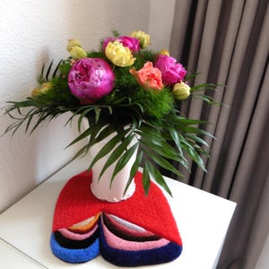Op de afbeelding: Een kleurrijk boeket van roze pioenrozen, gele tulpen en oranje bloemen in een witte vaas. De vaas staat op een rood tapijt met blauwe, zwarte en roze patronen. De opstelling staat op een witte tafel.