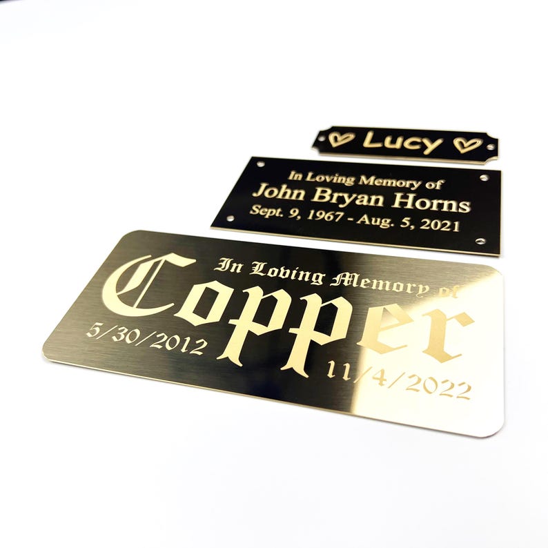 May include: A collection of memorial plaques in various shapes and sizes. The plaques are gold and black, with engraved text including names, dates, and the phrase "In Loving Memory." One plaque reads "Lucy" with hearts.