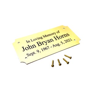 May include: A rectangular gold-colored memorial plaque with rounded corners and four screw holes. The plaque reads "In Loving Memory of John Bryan Horns Sept. 9, 1967 - Aug. 5, 2021." Four gold screws are included.