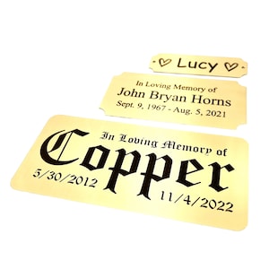 May include: Three gold-colored memorial plaques with black text. The largest plaque reads "In Loving Memory of Copper" with dates. The middle plaque says "In Loving Memory of John Bryan Horns." The smallest plaque says "Lucy."