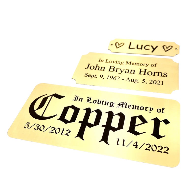 May include: Three gold-colored memorial plaques with black text. The largest plaque reads "In Loving Memory of Copper" with dates. The middle plaque says "In Loving Memory of John Bryan Horns." The smallest plaque says "Lucy."
