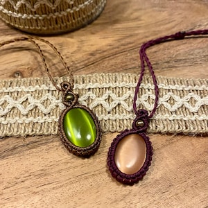 Op de afbeelding: Twee handgemaakte macramé kettingen. Eén heeft een lichtgroene ovale edelsteen, de andere een perzik-kleurige ovale edelsteen. Beide zijn gezet in een geweven bruine macramé setting en hangen aan een bijpassend koord.
