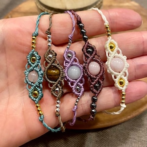 Puede incluir: Cinco pulseras de macramé con cordones de diferentes colores y cuentas de piedras preciosas. Las pulseras presentan una variedad de piedras, incluyendo cuarzo rosa, ojo de tigre y una piedra azul claro.