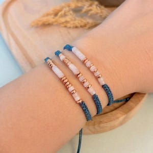 Può includere: Tre braccialetti fatti a mano con un cordino blu intrecciato e una combinazione di perline bianche, rosa e dorate. I braccialetti sono esposti su un polso, evidenziando il design delicato e la palette di colori.