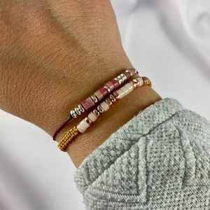 Puede incluir: Dos pulseras de cuentas. Una pulsera presenta cuentas rosas y plateadas, la otra tiene cuentas blancas y oro rosa. Ambas pulseras están en una muñeca, con un cordón amarillo tejido y un cordón morado oscuro.