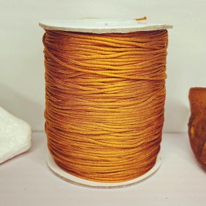 May include: A spool of gold colored cord or string. The cord is wound tightly around a white cardboard spool.
