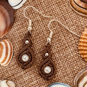 Pode incluir: Um par de brincos de macramé castanhos com um design em forma de gota, com uma pérola branca no centro e uma pequena conta prateada por cima. Os brincos têm ganchos de orelha prateados. Apresentados numa superfície castanha texturizada com conchas.