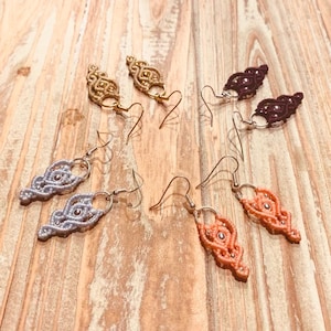 Peut inclure: Plusieurs boucles d'oreilles macramé faites à la main, de couleurs or, violet, argent et corail. Chaque boucle d'oreille présente un design tissé unique avec de petits accents métalliques et une fermeture à crochet. Les boucles d'oreilles sont présentées sur une surface en bois.