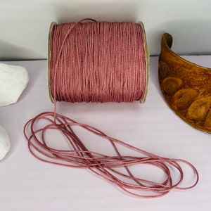 May include: A spool of dusty rose colored cord, with a portion of the cord unwound and lying on a white surface.
