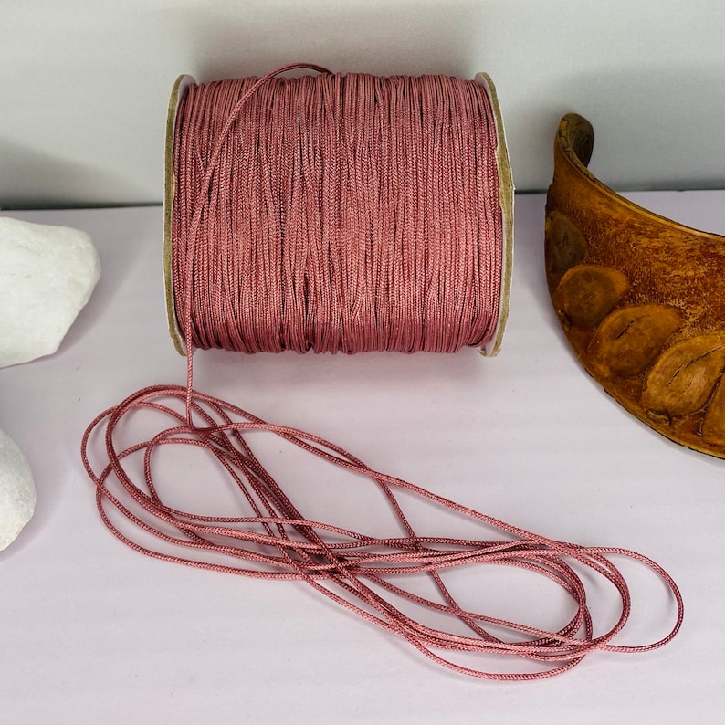 May include: A spool of dusty rose colored cord, with a portion of the cord unwound and lying on a white surface.