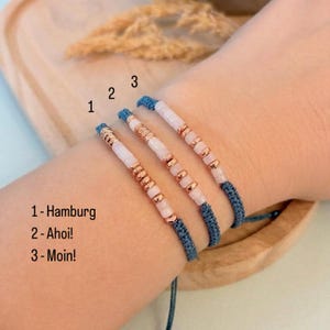 Könnte beinhalten: Drei Armbänder mit roségoldenen Akzenten und weißen und rosa Perlen. Die Armbänder haben ein blau gewebtes Band und sind mit dem Text "1- Hamburg", "2- Ahoi!" und "3- Moin!" beschriftet.