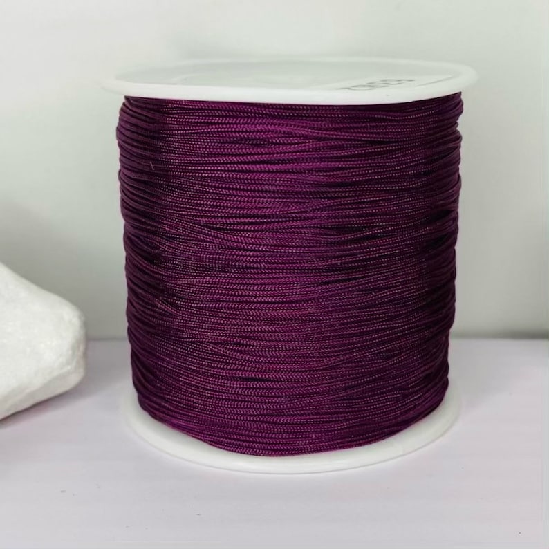 May include: A spool of deep purple cord, wound tightly on a white plastic bobbin. The cord is smooth and shiny, with a slight sheen.