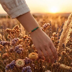 Könnte beinhalten: Ein Armband mit einer Reihe kleiner, zylindrischer grüner Perlen, die mit metallisch dunkelgrauen Zwischenstücken durchsetzt sind und von einer dünnen, geflochtenen goldenen Kordel gehalten werden. Das Design ist schlicht und linear und zeigt einen Kontrast zwischen den leuchtend grünen und den metallisch grauen Elementen auf der goldenen Kordel.