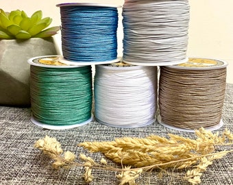 Pode incluir: Cinco carretéis de cordão de algodão encerado em diferentes cores: azul claro, cinza, verde, branco e marrom. Os carretéis estão dispostos em forma de pirâmide sobre uma superfície cinza texturizada.