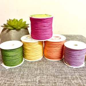 Op de afbeelding: Vijf spoelen met gekleurde draad, in groen, geel, oranje, paars en roze, zijn gerangschikt op een grijs, getextureerd oppervlak. De spoelen zijn gemaakt van wit plastic en hebben een label aan de zijkant.