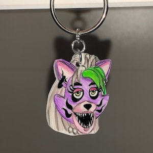 Puede incluir: Un personaje de dibujos animados morado y verde con una boca grande y dientes afilados. El personaje está unido a un llavero plateado.