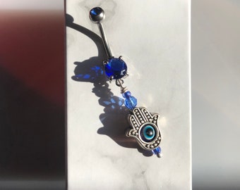 May include: A silver belly button ring with a blue crystal charm and a hamsa hand charm with a blue evil eye.
