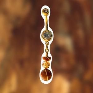 Pode incluir: Anel de umbigo dourado com um cristal transparente e uma pedra marrom pendurada com fio de ouro enrolado.