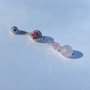 May include: A silver belly button ring with a pink gemstone and a dangling strand of pink beads and a large rose quartz bead. The jewelry is designed for body piercing.