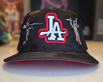 Pode incluir: Boné de beisebol camuflado preto com aba vermelha e as letras "LA" em branco com contorno vermelho. O boné apresenta figuras bordadas em cada lado.