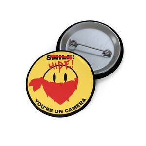 May include: A yellow button with a smiley face wearing a red bandana. The smiley face has the word "HIDE!" written over the word "SMILE!" and the text "YOU'RE ON CAMERA" around the bottom.