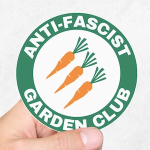 May include: A green and white round sticker with the text "Anti-Fascist Garden Club" and three orange carrots with green tops.