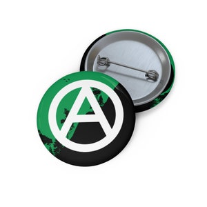 May include: A green and black button with a white anarchy symbol. The symbol is a circle with an A inside. The background is a green forest scene with black silhouettes of trees.