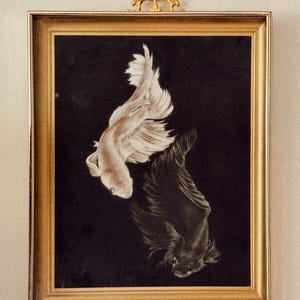 May include: A framed artwork featuring a white betta fish and a black betta fish swimming against a black background. The rectangular frame is gold with a decorative gold ornament at the top. The text "studio eleni" is printed at the bottom.