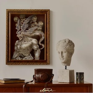May include: A framed rectangular wall art piece featuring a muscular, winged figure with a dark background, positioned next to a stone-colored bust of a human head on a pedestal, a brown leather camera case, a black pen, a book, and a small tin.