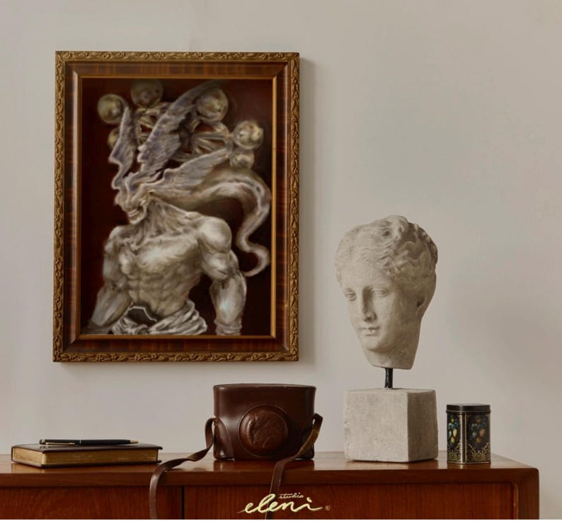 May include: A framed rectangular wall art piece featuring a muscular, winged figure with a dark background, positioned next to a stone-colored bust of a human head on a pedestal, a brown leather camera case, a black pen, a book, and a small tin.