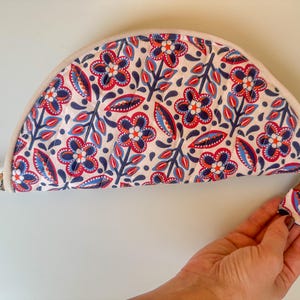 May include: A half-moon shaped floral print pouch with a cream-colored border and gold zipper. The pouch features a repeating pattern of red and blue flowers and leaves on a white background. The zipper pull is made of the same floral fabric.