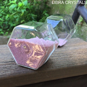 May include: Two clear glass geometric containers with hexagonal facets, each partially filled with fine, light purple granular material. The text LIBRA CRYSTALS is visible in the upper right corner of the image, set against a blurred green background.