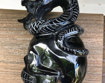 May include: A polished black stone carving featuring a coiled snake resting atop a human skull. The snake has detailed etched scales and an open mouth, while the skull displays defined eye sockets and teeth. The text LIBRA CRYSTALS is visible.