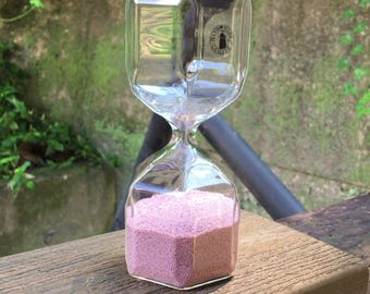 May include: A clear glass hourglass with a hexagonal shape containing fine pink sand. The top bulb features a small circular sticker with the text HAND-MADE QUALITY. The text LIBRA CRYSTALS is visible in the upper right corner of the image.