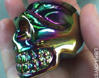 May include: A small, polished skull figurine featuring a reflective, iridescent finish that shifts between shades of purple, green, and gold. The surface is smooth and metallic, highlighting the anatomical details of the eye sockets and teeth. LIBRA CRYSTALS.