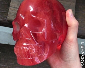 May include: A polished, translucent red skull carving with visible internal marbling and natural veining. The object features defined eye sockets, a nasal cavity, and detailed teeth. The text LIBRA CRYSTALS is printed vertically on the right side.