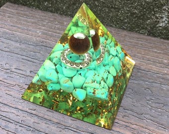 May include: A pyramid-shaped resin sculpture with turquoise stones and gold flakes. The sculpture has a gold-colored top and base. A brown and silver ring and a brown sphere are on top. The text "LIBRA CRYSTALS" is at the top.
