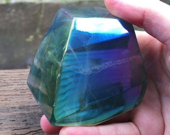May include: A faceted, iridescent crystal with a polished surface displaying deep blue, purple, and teal hues. The stone has a geometric shape with multiple flat faces and internal fractures. The text LIBRA CRYSTALS is visible.