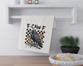 May include: A cream-colored kitchen towel featuring a black crow perched on a branch, surrounded by yellow daisies and a black and white checkered square background. The text F-CAW-F is printed in black above the crow illustration.