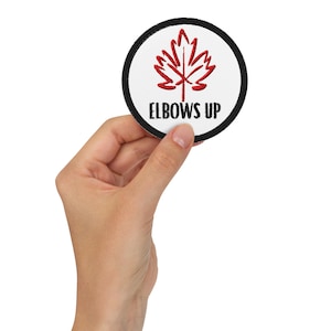 Puede incluir: Un parche redondo blanco con un borde negro. El parche tiene una hoja de arce roja bordada y el texto "ELBOWS UP" en negro.