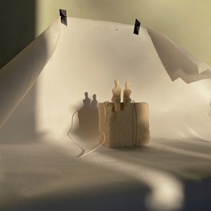 May include: A small, beige sculpture of two figures seated on a textured, square block. The sculpture is lit by warm light, casting shadows on the white backdrop. A thin string is attached to the block.