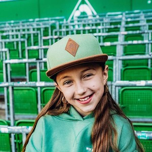 Op de afbeelding: Een lichtgroene honkbalpet met een bruine klep en een ruitvormige bruine patch aan de voorzijde, gecombineerd met een bijpassende lichtgroene hoodie met de witte tekst WIE B op de borst, gedragen door een glimlachend persoon.