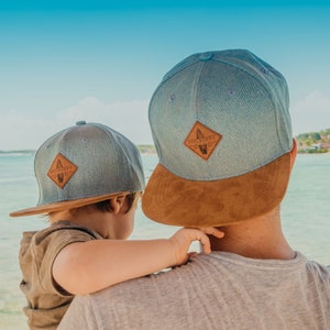 Könnte beinhalten: Ein Vater und sein Sohn tragen passende Baseballkappen aus blauem Denim und braunem Wildleder. Die Kappen haben einen braunen Leder-Patch mit dem Text "Soulbuddy" und einer Surfboard-Grafik.