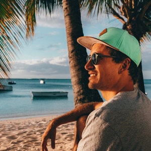 Könnte beinhalten: Eine Person mit einer grünen und beigen Baseballkappe und Sonnenbrille, die zum Meer lächelt. Die Person sitzt an einem Sandstrand mit Palmen und Booten im Hintergrund.