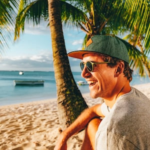 Könnte beinhalten: Ein Mann mit einer grün-braunen Baseballkappe und Sonnenbrille lächelt, während er an einem Strand mit einer Palme im Hintergrund sitzt.