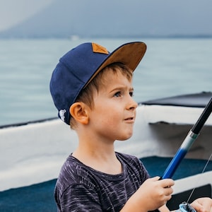 Könnte beinhalten: Ein kleiner Junge trägt eine blaue und braune Baseballkappe mit einem weißen Logo und hält eine Angelrute. Er schaut nach oben und scheint sich auf etwas in der Ferne zu konzentrieren.
